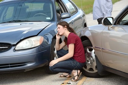 A woman on the phone crouching between two cars who have crashed into each other.