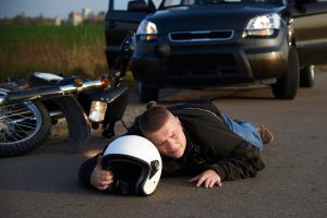 A motorcyclist lays on the road with their helmet in their hands. A car is behind them. 