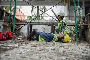 An injured worker lying on the floor next to scaffolding, with a colleague knelt next to them and pointing upwards.