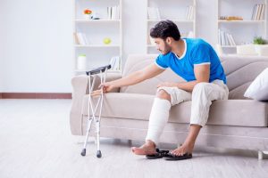 A man with one leg in a cast, sitting on a sofa and reaching to his right for a pair of crutches.