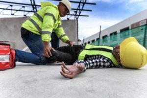 A builder in a high-visibility vest and hard hat tends to another who is injured on the floor of a construction site.