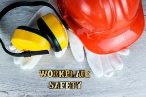 A red helmet, yellow headphones, and white gloves on a table with 'workplace safety' written out in wooden letters.