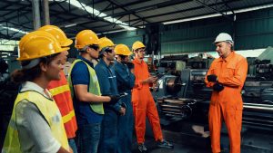 A factory manager trains younger staff in safe working methods while they all wear high-visibility clothing.