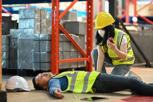 Man in high-vis lies on floor after injury at work. Woman crouches beside him and speaks into walkie talkie